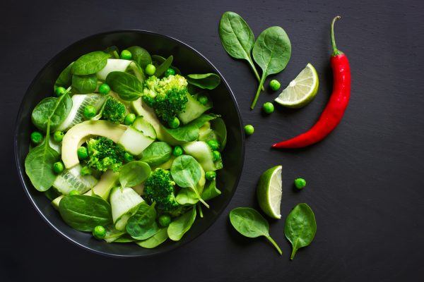 Skål med friske grønne grøntsager, avokado, broccoli mm. skålen er placeret på sort bord med limebåde og grønne blade, en rød chili giver kontrast